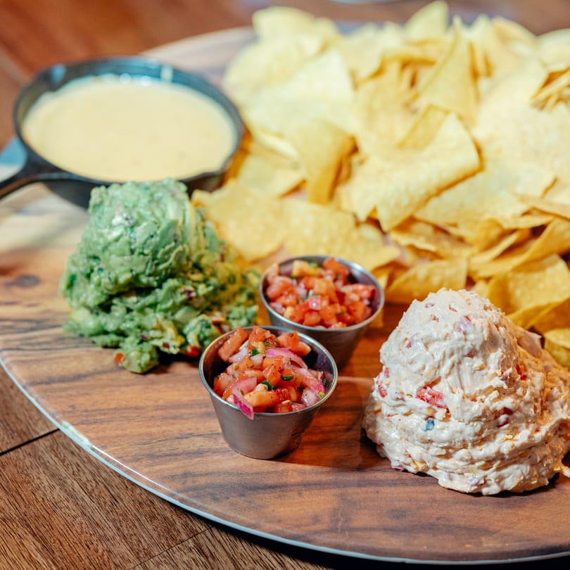 A wooden board with tortilla chips, queso, guacamole, pico de gallo, and a creamy dip.