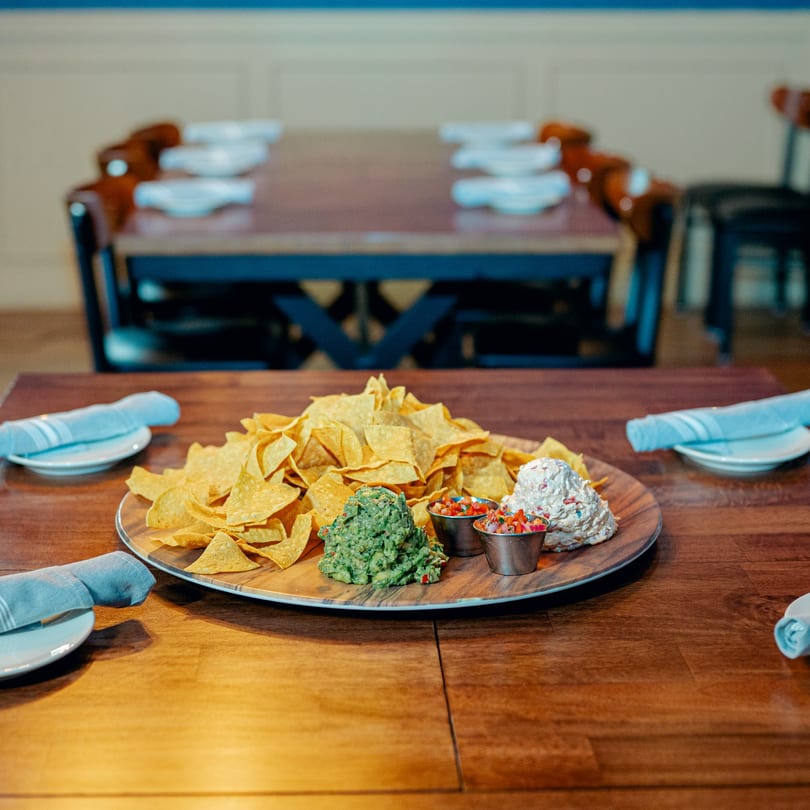 A wooden platter of tortilla chips, guacamole, salsa, and a creamy dip on a tavern table.