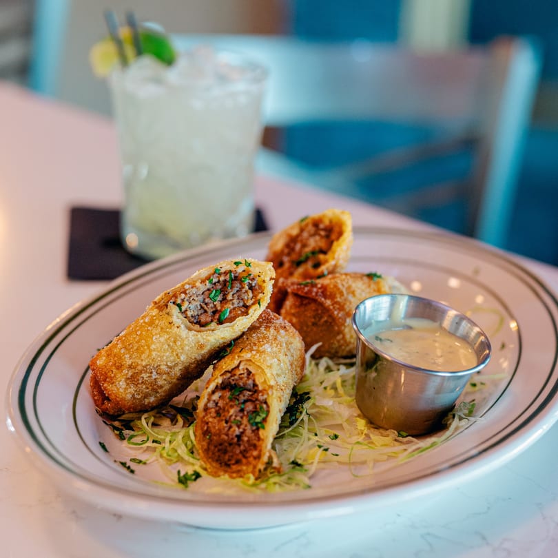 Four crispy egg rolls, one cut open, with a side of dipping sauce and a cocktail.