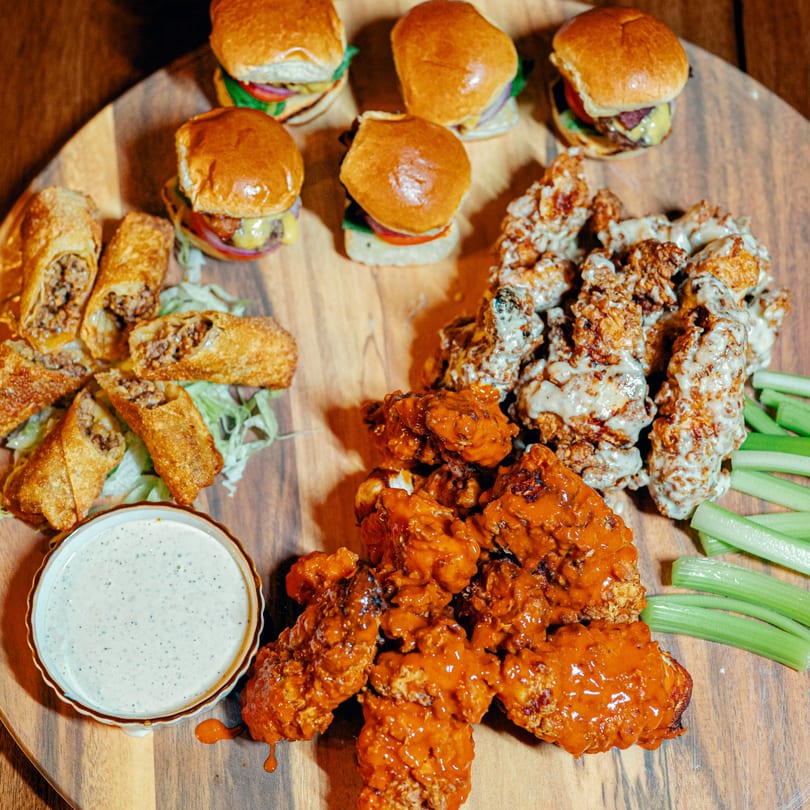 A wooden board with sliders, egg rolls, buffalo wings, garlic parmesan wings, celery, and ranch dip.
