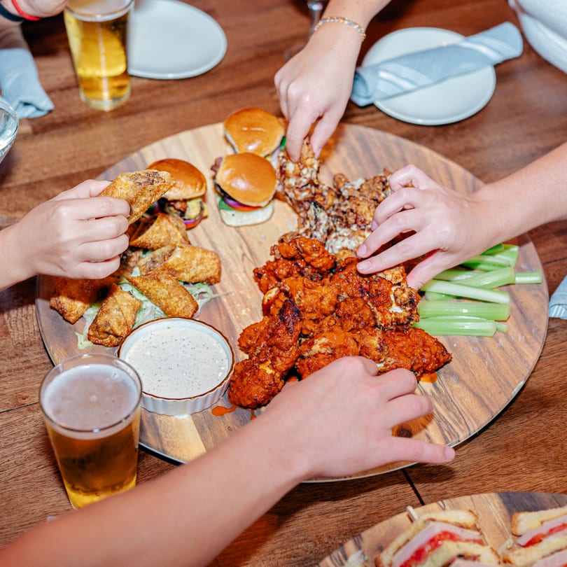 Hands reaching for sliders, wings, egg rolls, and cauliflower bites on a large wooden platter.