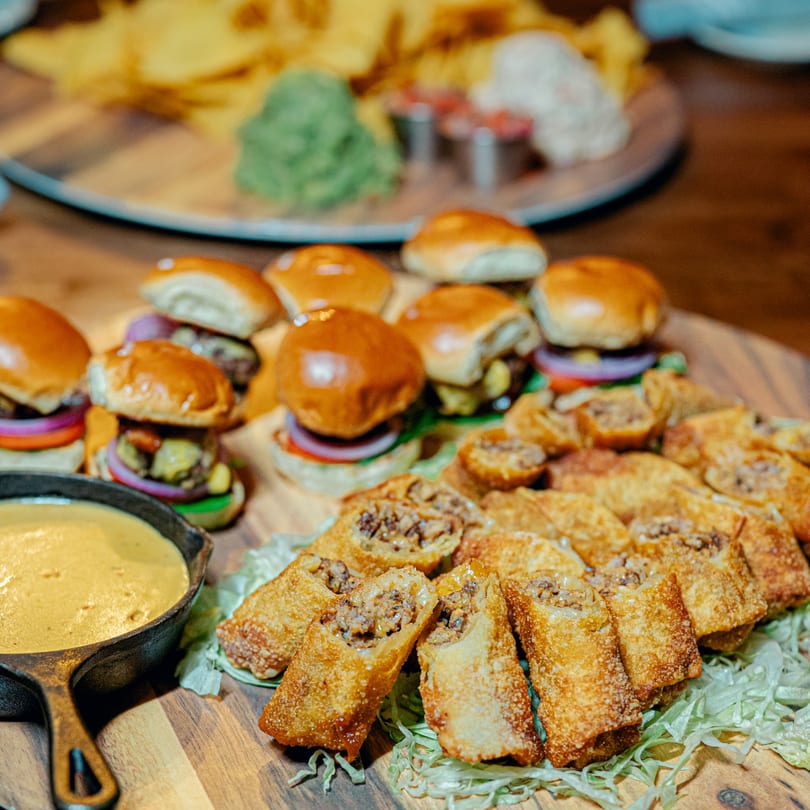 A wooden board with sliders, egg rolls, and a cast iron skillet of cheese dip.