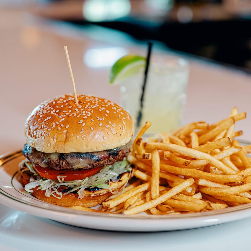 83 Tavern's burger with melted cheese, lettuce, tomato, and a side of golden fries and a drink.